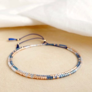 May include: A delicate beaded bracelet with a layered design featuring blue, white, and gold beads. The bracelet is adjustable with a sliding knot and a small tassel.