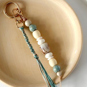 May include: A handcrafted keyring with a gold-tone clasp and ring. The keyring features a strand of wooden beads in cream, beige, and seafoam green, plus a braided teal and beige tassel. The beads include a shell-shaped accent.