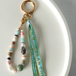 May include: A gold-coloured keyring with a beaded charm and a teal ribbon. The charm features a variety of beads in pastel colours, seashells, and a ceramic bead. The teal ribbon has gold text that reads "OCEAN CHILD".