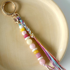 May include: A beaded keychain with a gold-coloured clasp. The keychain has a braided section and a strand of pink, yellow, and white beads. A tassel of multicoloured threads hangs from the bottom.