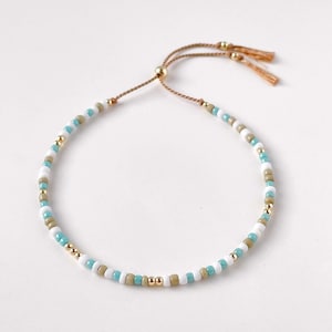 May include: A delicate beaded bracelet with a sliding knot closure. The bracelet features small, round beads in shades of white, light blue, and gold.