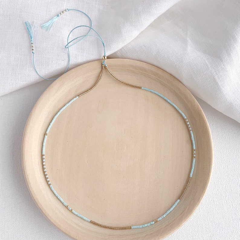 Minimalist blue and gold beaded necklace on a silk string with a slider clasp in 14c gold fill with tassel ends on a wooden plate