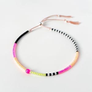 May include: A vibrant beaded bracelet with a sliding knot fastening. The bracelet showcases a lively mix of colours, including black, pink, yellow, and orange beads. The adjustable cord is a light pink, and the bracelet is set against a white backdrop.