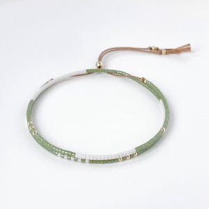 May include: A minimalist beaded bracelet in olive green and white, interspersed with gold-coloured beads. It has an adjustable brown cord fastening with gold details. The bracelet's design is simple and elegant, suitable for everyday wear, with a circumference of approximately 18 cm.