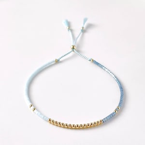 May include: A delicate blue and gold beaded bracelet with a sliding knot closure. The bracelet features a gradient of light blue beads and a row of gold beads.