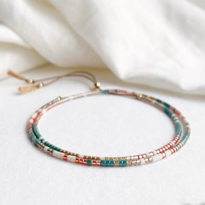 May include: A delicate double-strand bracelet featuring a mix of teal, white and rose gold beads. The bracelet has a sliding knot closure and a tassel.