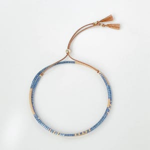 May include: Delicate adjustable bracelet. The bracelet is made of small, light blue and beige beads. It has a brown cord with a sliding knot for adjustment and small tassels. The bracelet is designed to fit various wrist sizes.
