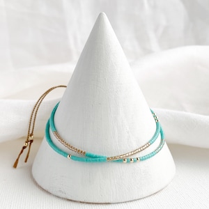 May include: Two layered bracelets, one turquoise blue and one gold, on a white cone shaped display stand.