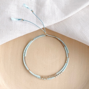 May include: A delicate double-strand bracelet with light blue and gold beads. The bracelet has a light blue cord with tassels at the ends.