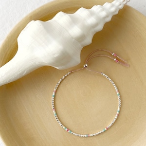 May include: A delicate silver bracelet featuring small white beads with colourful accents in pink and blue. The bracelet has a pink cord with a sliding knot for adjusting the fit.