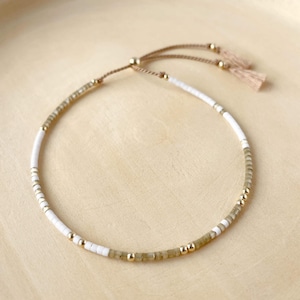 May include: A delicate white and green beaded bracelet with gold accents and a brown tassel. The bracelet is adjustable with a sliding knot.