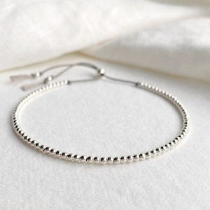 May include: A silver beaded bracelet with a sliding knot closure. The bracelet is made of small, round beads and has a simple, elegant design.