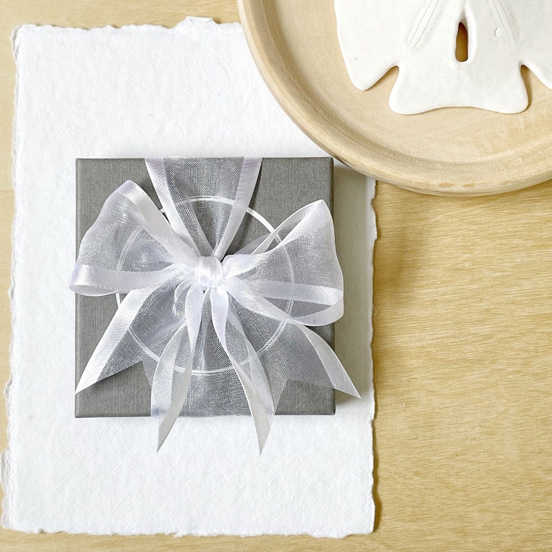 Grey jewellery gift box with white organza ribbon box sitting on a backdrop of white paper and wood