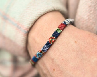 Peut inclure: Un bracelet tissé coloré avec des fils rouges, bleus, verts et blancs. Le bracelet est au poignet d'une personne.