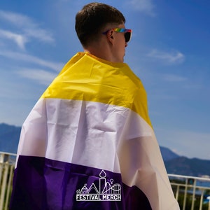 Könnte beinhalten: Eine Person trägt eine Regenbogen-Sonnenbrille und hält eine gelbe, weiße und violette Non-Binary-Pride-Flagge. Die Flagge hat den Text "Festival Merch" unten aufgedruckt.
