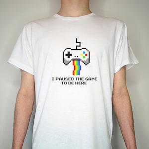 Può includere: Maglietta bianca con un'immagine pixelata di un controller per videogiochi con un arcobaleno che esce dal controller. Il testo "Ho messo in pausa il gioco per essere qui" è stampato sotto l'immagine.