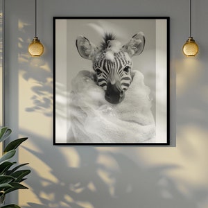 Op de afbeelding: Zwart-wit ingelijste print van een zebra veulen. De kop en gestreepte gezicht van de zebra zijn zichtbaar, gewikkeld in een zachte witte deken. De print hangt aan een grijze muur, met planten en een raam op de achtergrond.