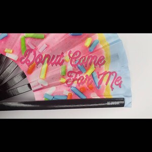 May include: A pink and blue hand fan with a donut design and the text "Donut Come For Me".