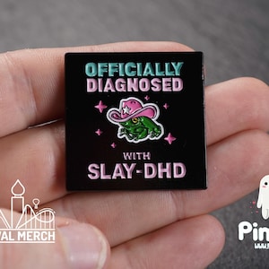 Puede incluir: Pin cuadrado negro con el texto "OFFICIALLY DIAGNOSED" en verde azulado y rosa. Una rana verde con un sombrero de vaquero rosa está en el centro. El texto "WITH SLAY-DHD" está debajo. Estrellas rosas rodean la rana.