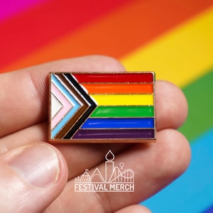 Op de afbeelding: Een gouden emaille pin met een regenboog pride-vlag met een zwart en bruin chevron-ontwerp. De pin wordt in een hand gehouden tegen een regenboog achtergrond.