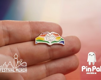 May include: A colorful enamel pin in the shape of hands forming a heart, with the word "love" in the center. The pin features a rainbow design and is held in a hand. The pin is a Festival Merch product.