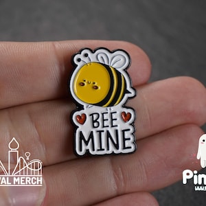 Op de afbeelding: Een schattige emaille pin met een gele en zwarte cartoon bij met een lachend gezicht. De pin heeft de tekst "BEE MINE" in witte letters met rode hartaccenten. De pin heeft een zwart-witte rand en een glanzende afwerking.