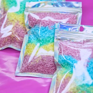 May include: Three clear plastic bags filled with colorful bath salts. The salts are layered in shades of pink, yellow, blue, and green. The bags are sealed with a zipper and have a holographic finish.