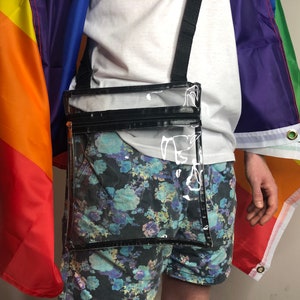 May include: A clear plastic crossbody bag with a black zipper and strap. The bag is worn over a person's shoulder and is in front of a rainbow flag.