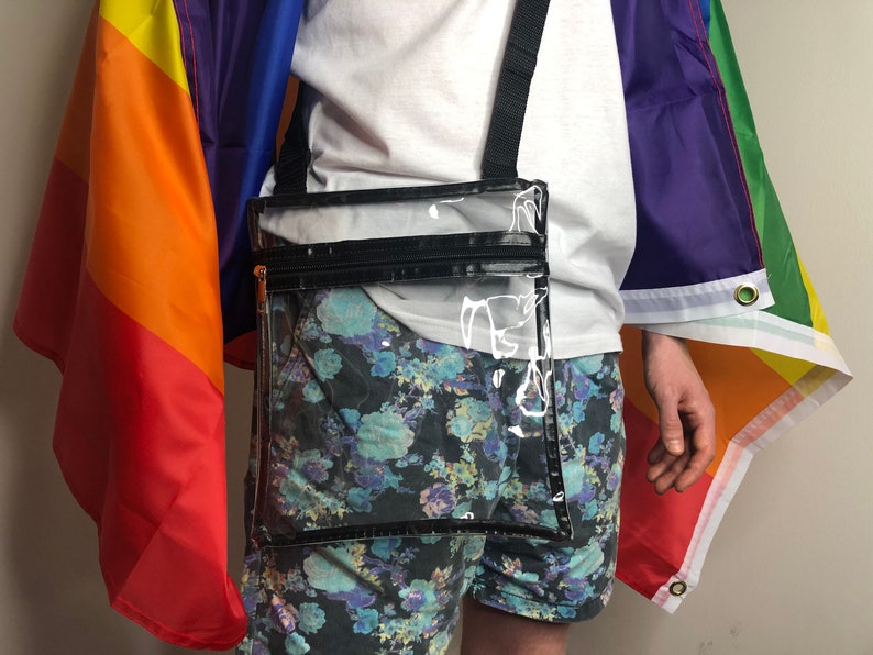 May include: A clear plastic crossbody bag with a black zipper and strap. The bag is worn over a person's shoulder and is in front of a rainbow flag.