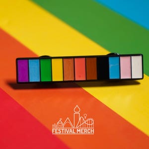 May include: A rectangular enamel pin with a rainbow color palette, including purple, blue, green, yellow, orange, and pink. The pin is set against a background of diagonal rainbow stripes. The Festival Merch logo is at the bottom.