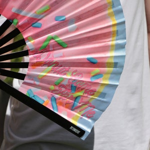 May include: A pink and blue hand fan with a donut design and the text "Donut Give Up On The Vibe". The fan is made of paper and has a black handle.