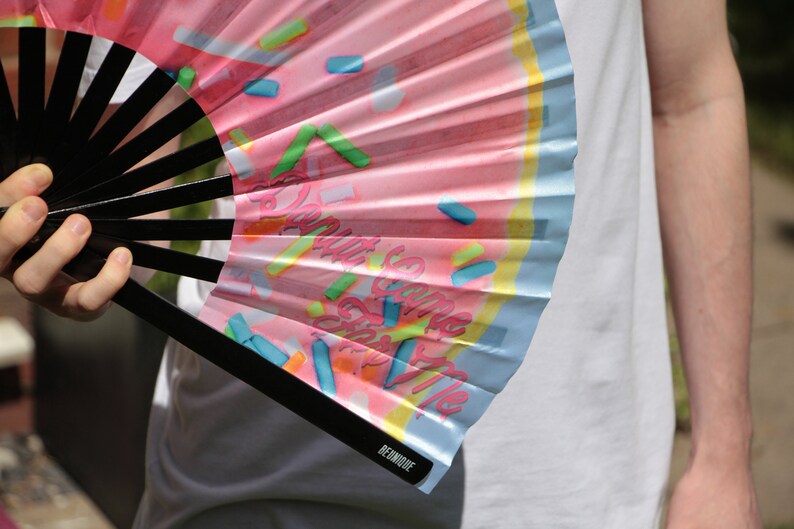 May include: A pink and blue hand fan with a donut design and the text "Donut Give Up On The Vibe". The fan is made of paper and has a black handle.