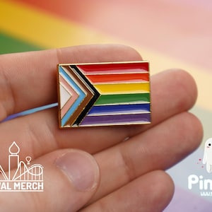 May include: A rectangular enamel pin with a gold border, displaying the Progress Pride flag design. The pin features the rainbow colours, the transgender flag colours, and black and brown stripes. The pin is held in a hand.