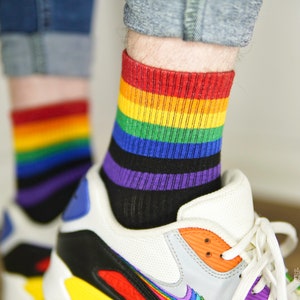 Op de afbeelding: Een paar witte sneakers met regenboogkleurige sokken. De sokken hebben rode, oranje, gele, groene, blauwe, indigo en paarse strepen.