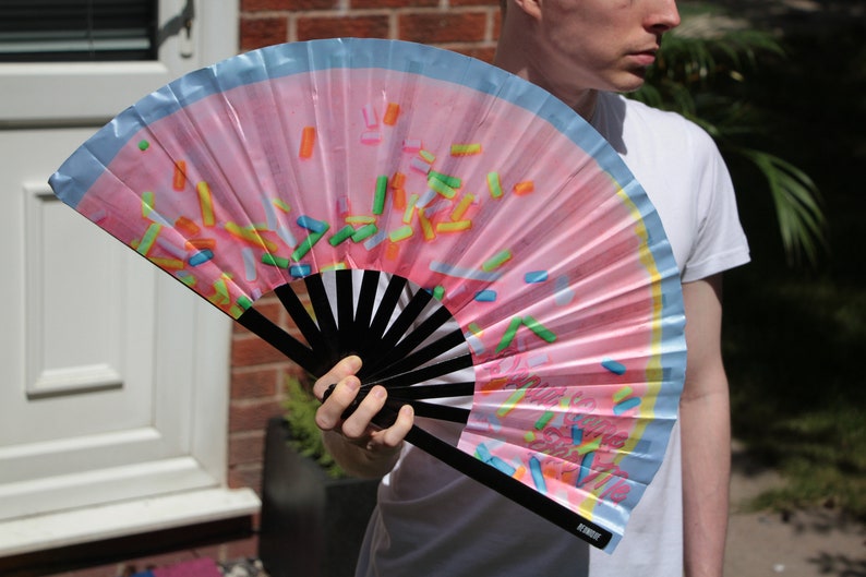 May include: A pink and blue hand fan with a sprinkle pattern. The fan is open and has a black handle with the word "remarkable" printed on it.