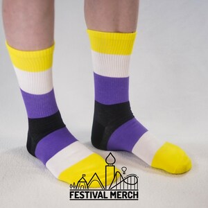 Puede incluir: Calcetines con los colores de la bandera del orgullo no binario. Los calcetines presentan rayas amarillas, blancas, moradas y negras. El logotipo de Festival Merch está en la parte inferior.
