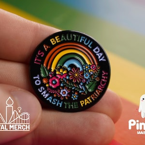 May include: A circular black enamel pin featuring a rainbow and floral design. The pin has the text "It's a beautiful day to smash the patriarchy." The pin is held in a hand, with the Festival Merch and Pin Pal logos visible.