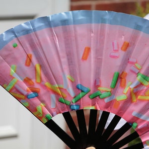 May include: A pink hand fan with a blue border and a pattern of colorful sprinkles. The fan has black wooden ribs.