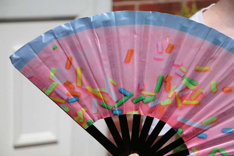 May include: A pink hand fan with a blue border and a pattern of colorful sprinkles. The fan has black wooden ribs.
