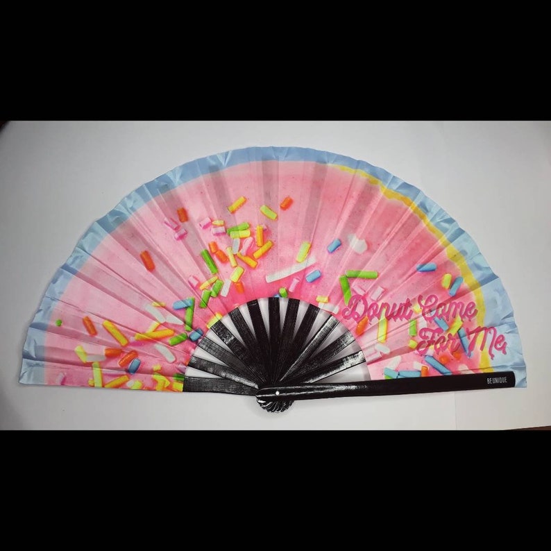 May include: A pink and blue hand fan with a donut design and the text "Donut Come For Me". The fan has black bamboo ribs and a black handle.