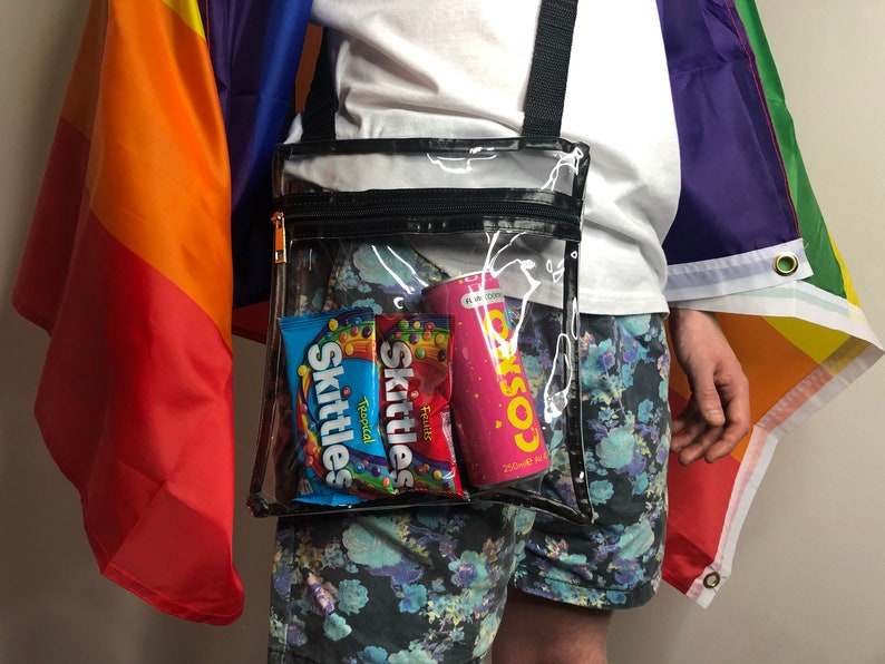 May include: A clear plastic crossbody bag with a black zipper and strap. The bag contains two packs of Skittles candy and a pink can of COSMO soda.