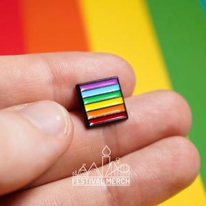 May include: A rainbow enamel pin with a black border. The pin has six stripes of colour: purple, blue, turquoise, green, yellow, and red.