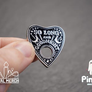 Op de afbeelding: Een hartvormige, zwart-zilveren emaille pin met de woorden "SO LONG AND GOODNIGHT" en maandetails. De pin heeft een gat in het midden en wordt tegen een grijze achtergrond gehouden. De pin is een novelty item.