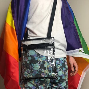 May include: A clear plastic crossbody bag with a black zipper and strap. The bag is worn over a white t-shirt and floral print shorts.