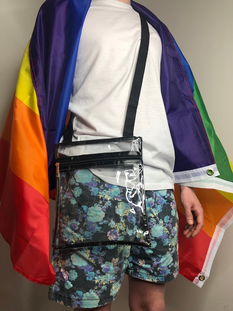 May include: A clear plastic crossbody bag with a black zipper and strap. The bag is worn over a white t-shirt and floral print shorts.