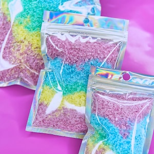 May include: Three iridescent bags filled with colorful bath salts. The salts are layered in pink, blue, and yellow.