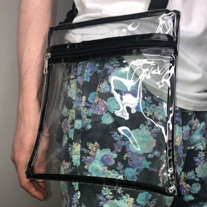 May include: A clear plastic crossbody bag with a black zipper and strap. The bag has a black trim around the edges.