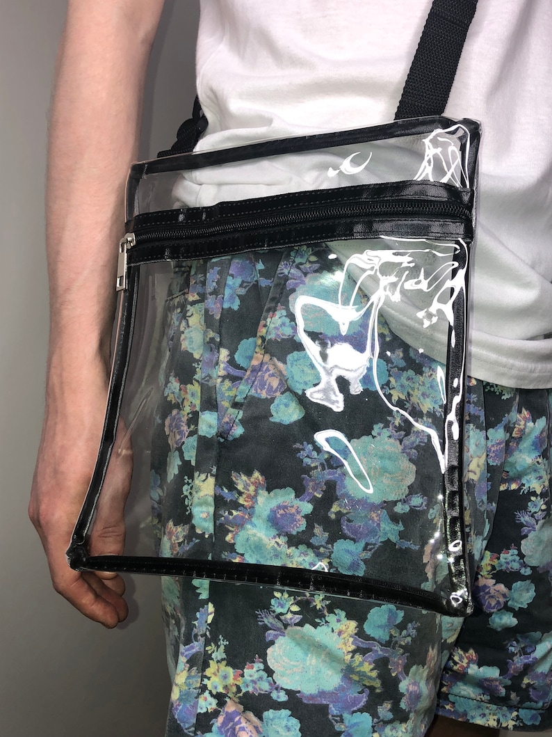 May include: A clear plastic crossbody bag with a black zipper and strap. The bag has a black trim around the edges.