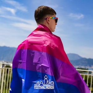 May include: A person wearing rainbow-coloured sunglasses and a large, colourful flag draped over their shoulders. The flag has stripes of pink, purple, blue, and white. The flag has the text "Festival Merch" printed on the bottom.