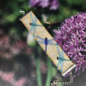 May include: A beaded bracelet with a light beige background and three blue and yellow dragonfly designs. The bracelet has a silver clasp and a silver charm with a dragonfly design.